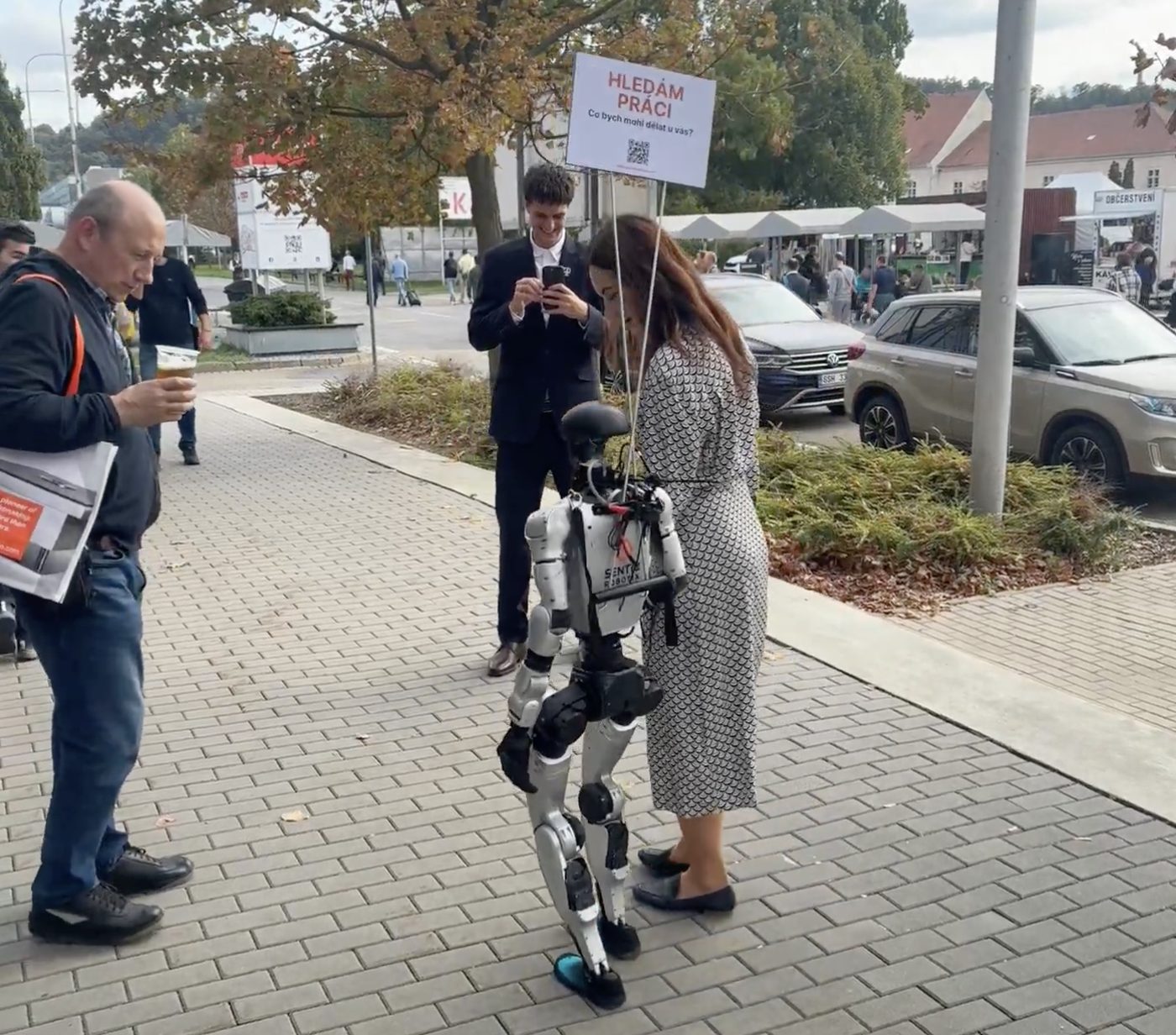 Senti s cedulí HLEDÁM PRÁCI, vedený za ruku před halami MSV